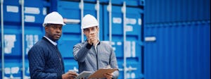 Two engineers in white hardhats carrying clipboards and a tablet stand in front of a blue shipping container
